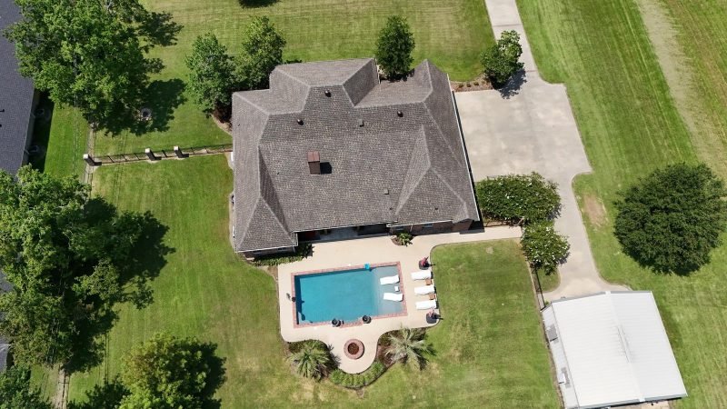 Straight-down drone view of a brown shingle hip roof, rectangular pool and long driveway