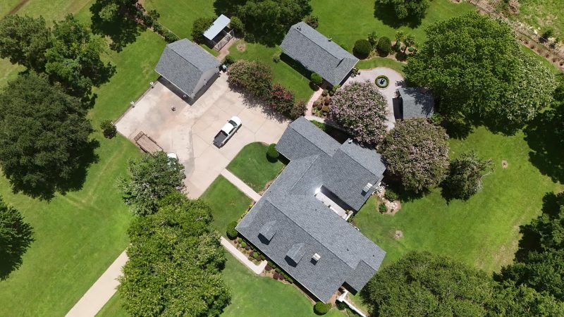 Drone view of a home and outbuildings with new gray shingle roofs among lawns and trees