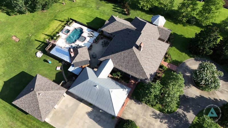 Drone view of a large home with shingle roof sections, a light metal garage roof and a pool courtyard