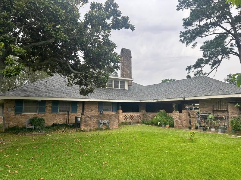 Long brick ranch home with a new gray roof and tall brick chimney, framed by oak branches