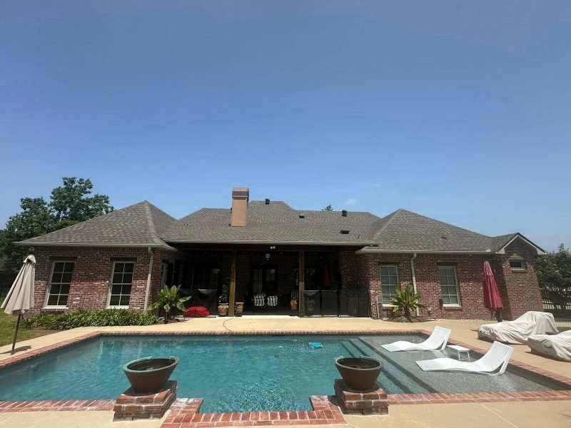 Rear of a brick home with a new brown roof over a covered patio, seen across the pool