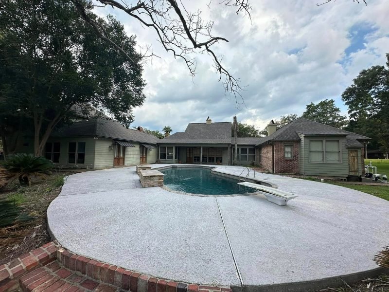 Curved pool with a diving board in front of a home with new gray roofs