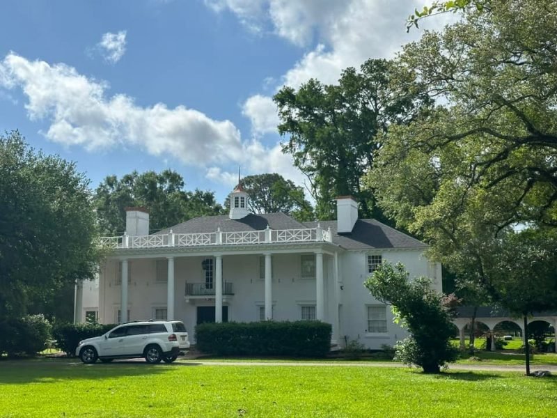 Large white two-story home with tall columns, a balcony and a cupola, under big oaks