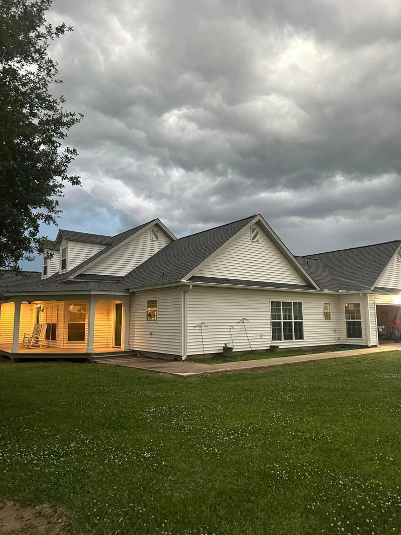 White-sided home with new dark roofs and a lit back porch at dusk