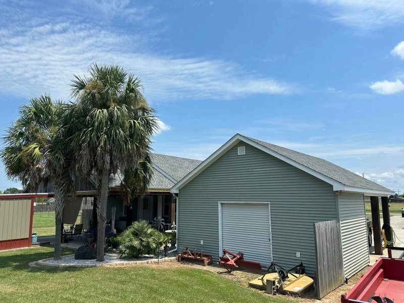 Green-sided garage building with a roll-up door and new shingle roof beside palm trees